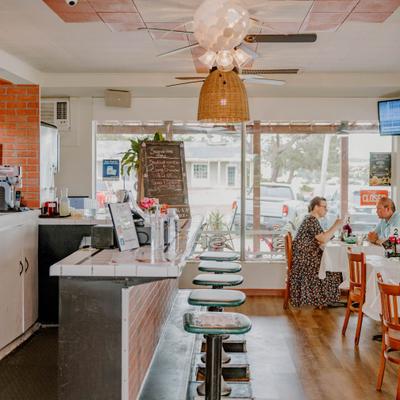 A bright, casual diner interior with bar stools, wooden tables, and patrons.