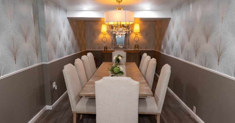 Dining room with a long table, upholstered chairs, chandelier and patterned wallpaper.