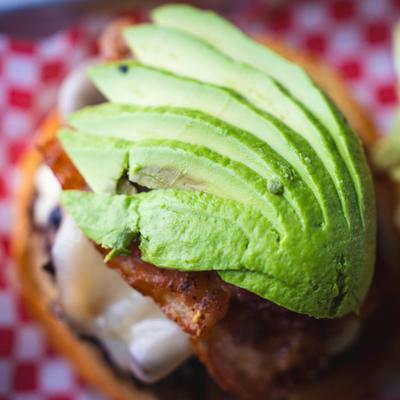 A close-up of a burger topped with sliced avocado and crispy bacon.