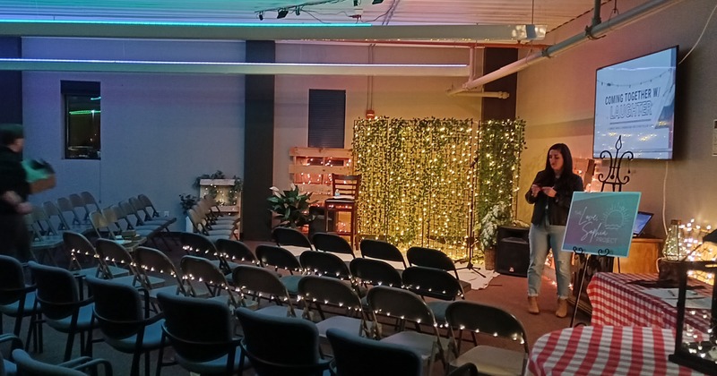 A woman performs in an event room in front of a row of chairs, while being illuminated by L.E.D. lights.