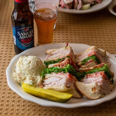 Turkey club sandwich served with potato salad, a pickle spear, and a beer.