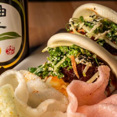 Buffalo chicken bao buns served alongside shrimp crackers and bottled drink.