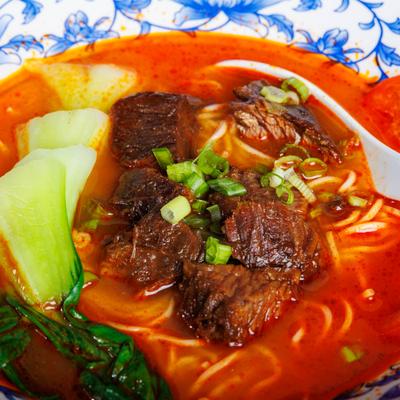 Close-up of spicy beef noodle soup with bok choy, tomatoes, and scallions.