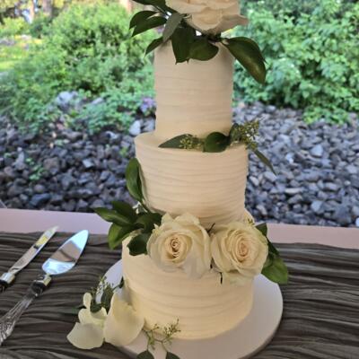 A white wedding cake with white roses and green leaves on a table, stone and greenery backdrop.
