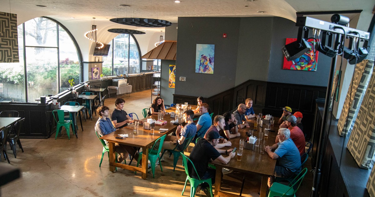 A diverse group of people sit at a long wooden table in a restaurant with large arched windows