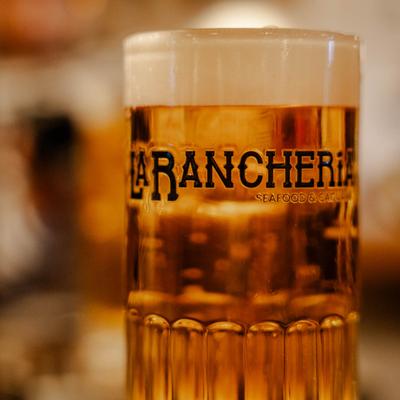 A tall glass labeled La Rancheria is filled with a foamy beer, close up.