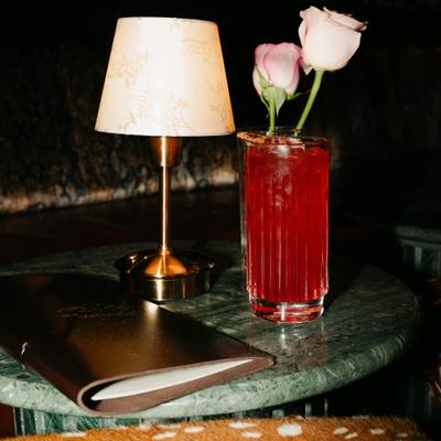 A red cocktail sits on a green marble table, with a menu, lamp, and roses.