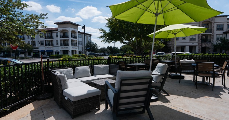 Outdoor patio with cushioned seating under green umbrellas, surrounded by greenery and buildings