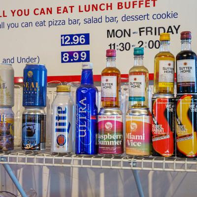 Canned and bottled drinks displayed on a shelf above a lunch buffet sign showing prices and hours.