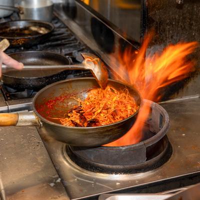 Chef stirring spicy noodles in a pan over a hot flame on a stove.