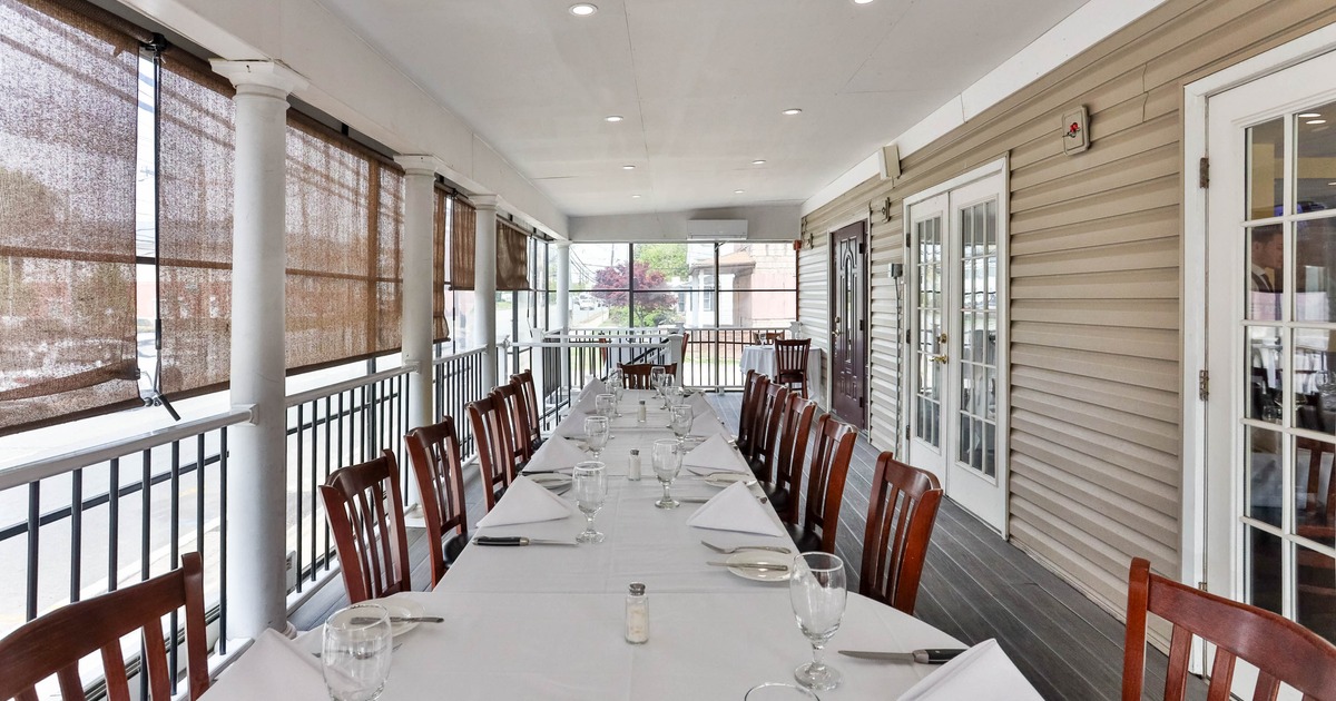 A long table with white tablecloth and place settings on an enclosed porch with wooden chairs