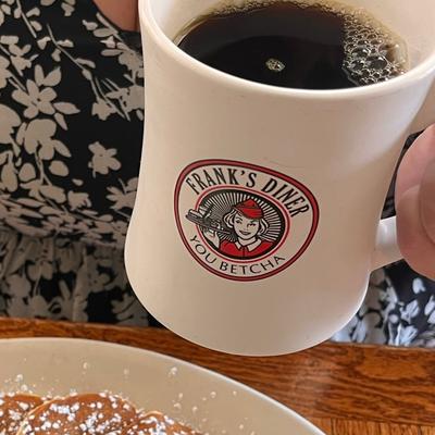 A white mug with Frank's Diner logo filled with coffee, held by a person.