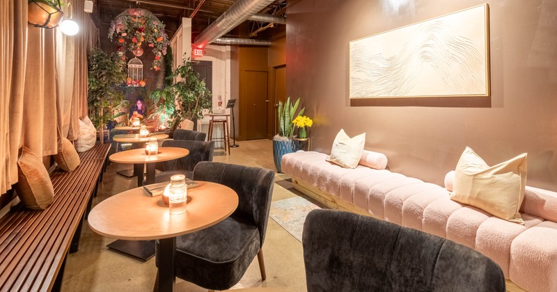 Cozy lounge with wooden tables, pink bench seating, and decorative plants under warm lighting