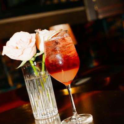 A tall glass with Aperol spritz sits next to two soft pink roses in a clear vase.