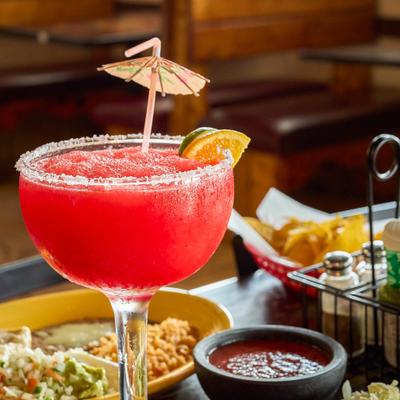 Frozen strawberry margarita with citrus and paper umbrella garnish surrounded by Mexican dishes.