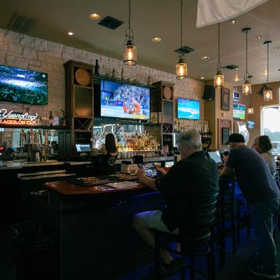 Bar interior with patrons, TVs playing sports, liquor bottles, and pendant lights.