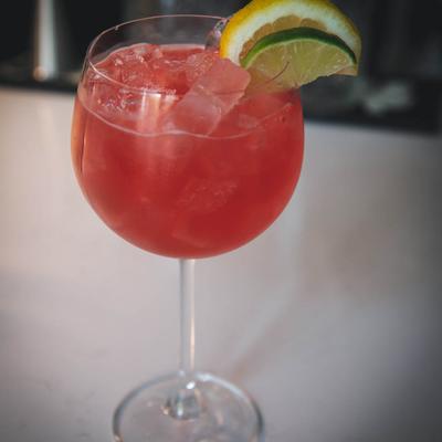 Red cocktail in a balloon glass, garnished with lime and lemon wedges.