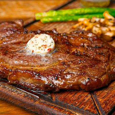 A grilled steak topped with herb butter on a wooden board with asparagus and mushrooms.