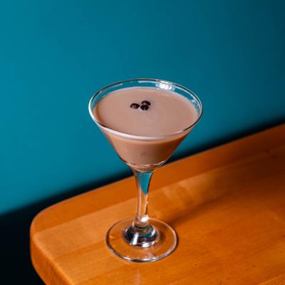 Espresso martini rests on a wooden table against a blue wall backdrop.