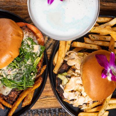 Two gourmet burgers with fries and a cocktail garnished with a purple orchid.