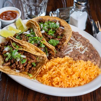 Beef tacos topped with cilantro and onions, served with rice, refried beans, and lime wedges.