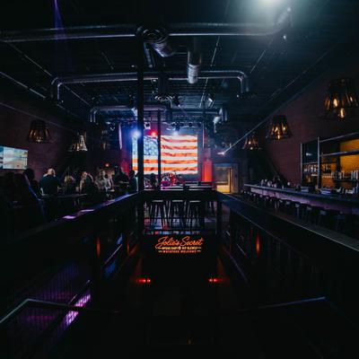 Interior featuring an illuminated American flag on the stage wall, overhead pipes, and a long bar.