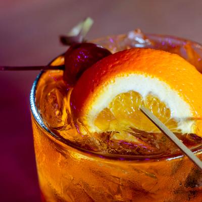 A glass of Old Fashioned garnished with a slice of orange and a cherry on a stick.