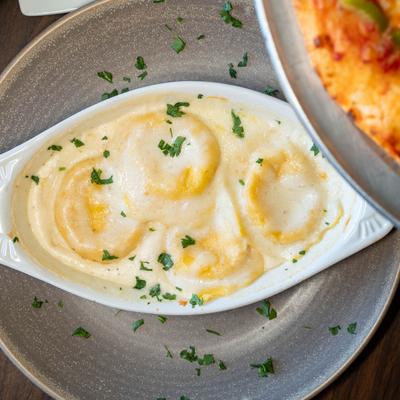 Ravioli pasta in a white, oval shaped dish on a gray plate on a table, a pizza is visible.