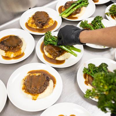 Food worker plating braised beef with broccolini on multiple prepared dishes.