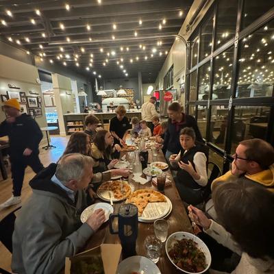 Group of people dining indoors around table with pizza and shared dishes.