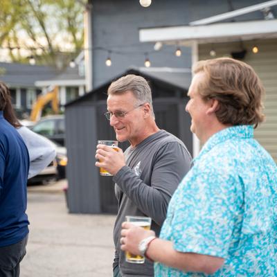 Two men holding drinks at an outdoor gathering with string lights and a shed in the background.