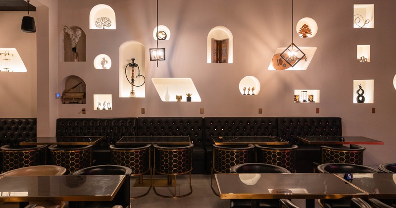 Interior of a stylish café featuring a light-colored wall with decorative alcoves, dark tufted seating, and gold-edged tables