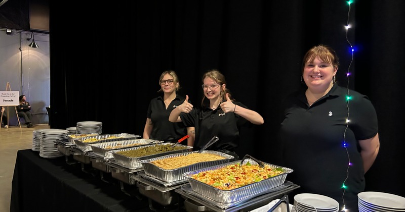 Three smiling servers stand behind a buffet table with various trays of food