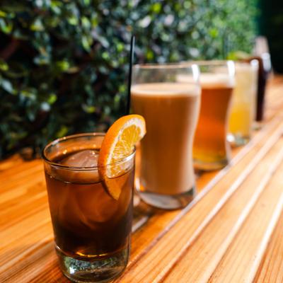 Old Fashioned Sailor and other cocktails lined up on a wooden table with a lush green background.