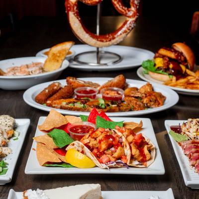 Various dishes on a table including fish tacos, chicken wings, pretzel, and burger with fries.
