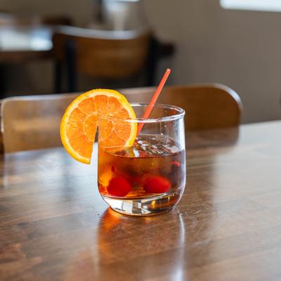 A glass of Old Fashioned with an orange slice, cherries, and a red straw, sits on a wooden table.
