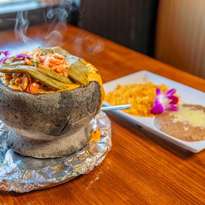 Steaming hot molcajete dish served with sides of rice and refried beans with melted cheese.