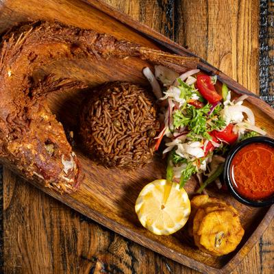 Whole fried fish with black rice, salad, plantains, red sauce, and lemon garnish.