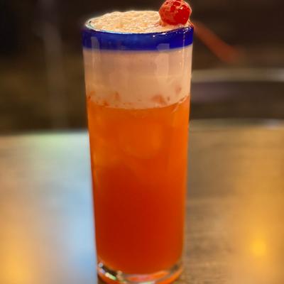 An orange cocktail in a blue-rimmed glass with cherry garnish.