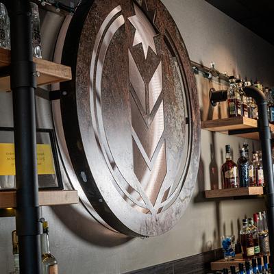 Large metal brewery logo on the wall above bar shelves.