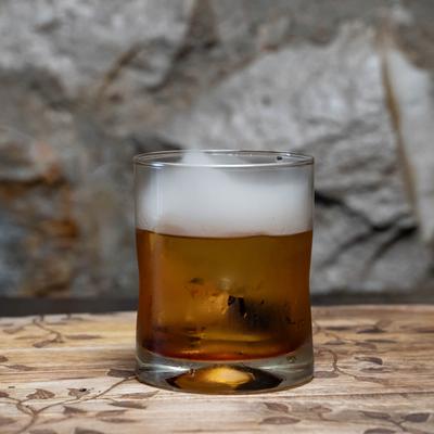 A smoked amber drink on top sits on a wooden table, a textured stone wall in the background.