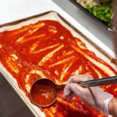 A food worker spreading tomato sauce on a pizza crust on a sheet pan.