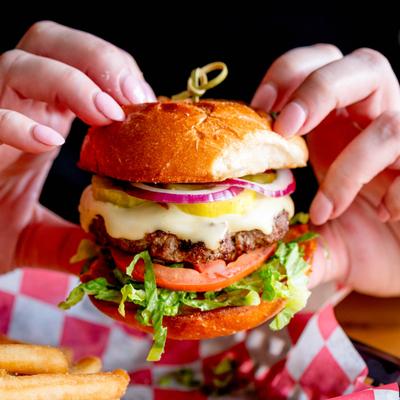 Hands holding a double cheeseburger with red onions, tomatoes, and lettuce.