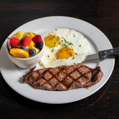 Steak and eggs served on a white plate with a bowl of fresh fruit.