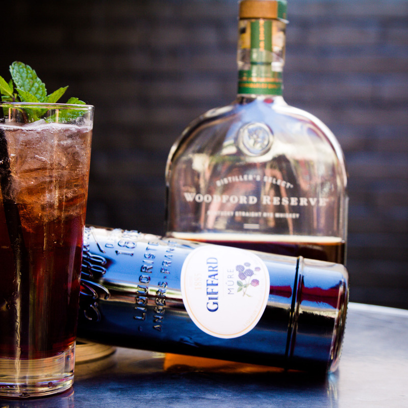 A dark cocktail and bottles of Giffard and Woodford Reserve