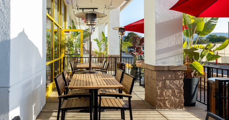 Outdoor patio with wooden tables, chairs, red umbrellas and lush plants under string lights