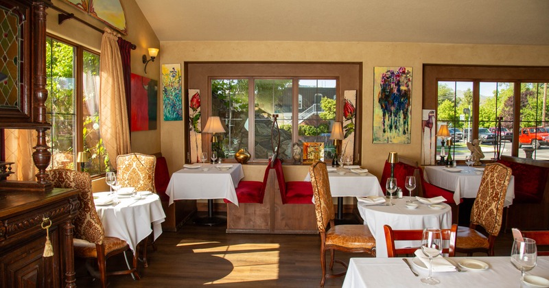 Restaurant interior with white tablecloths, ornate chairs, abstract paintings, and large windows with outdoor views