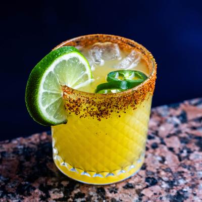 Yellow cocktail with lime slice, sliced jalapenos, and a chili rim.