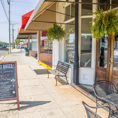 Sidewalk with storefront, awning, hanging plants, chalkboard and a dark metal bench.