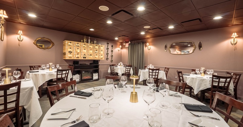 Interior of a warmly lit dining room with white tablecloths, wine glasses and decorative mirrors.